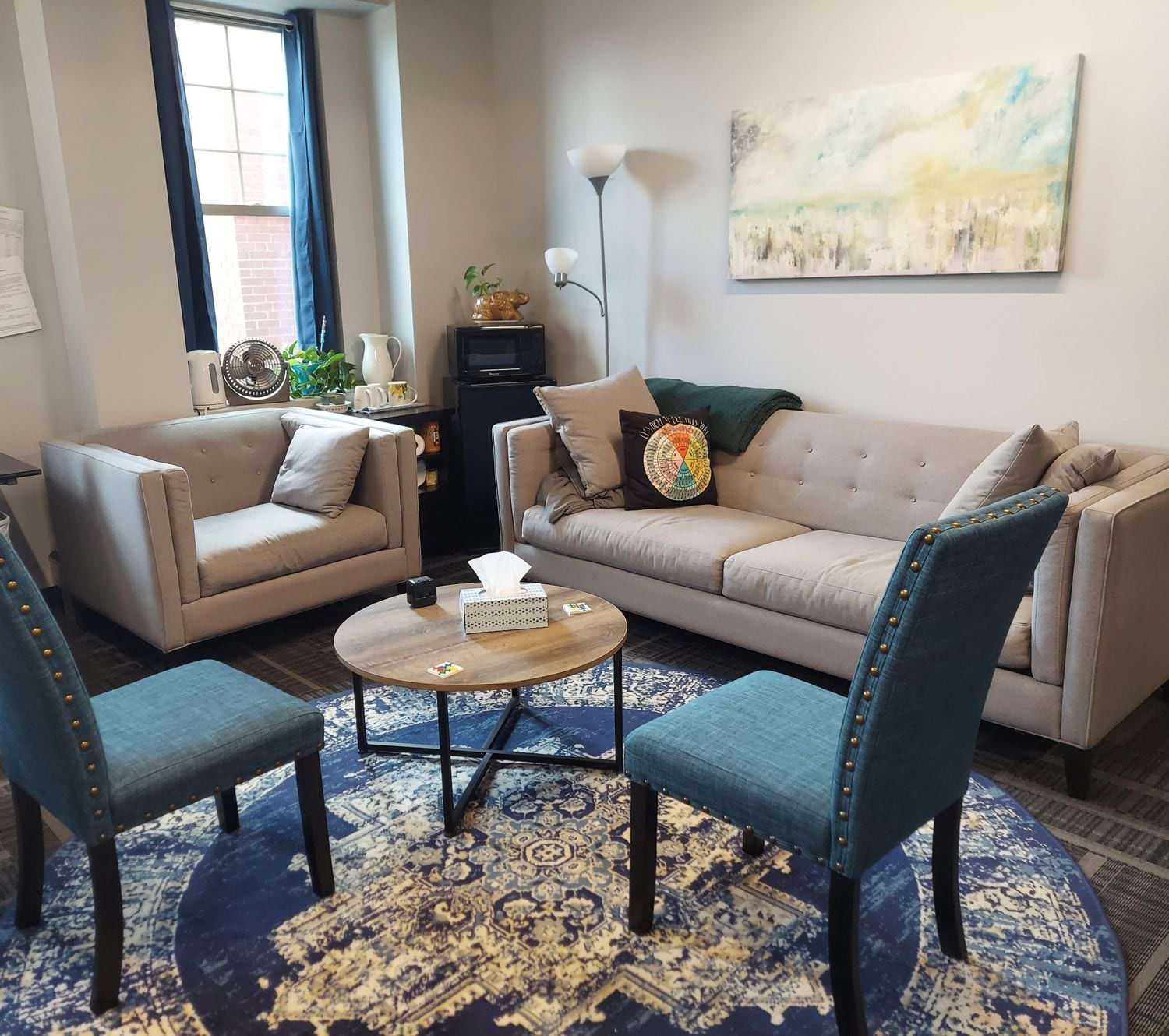 Inside the Dormont office — a beige couch, blue accent chairs around a round wooden coffee table, soft natural light from a tall window, and a landscape painting on the wall