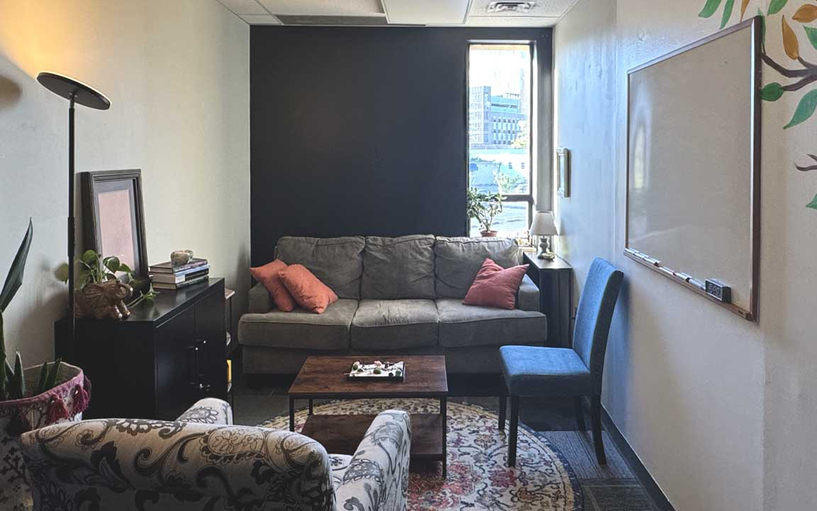 Inside the East Liberty office — a gray couch with coral pillows along a dark accent wall, a paisley armchair, and a city view through the window
